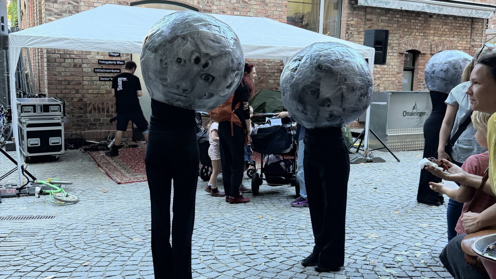 Drei Kugelköpfe im WUK-Hof: schwarz gekleidete Performerinnen, deren Köpfe große graue Kugeln aus Foto-Collage sind, stehen auf dem Kopfsteinpflaster zwischen Ziegelbauten und Publikum.
